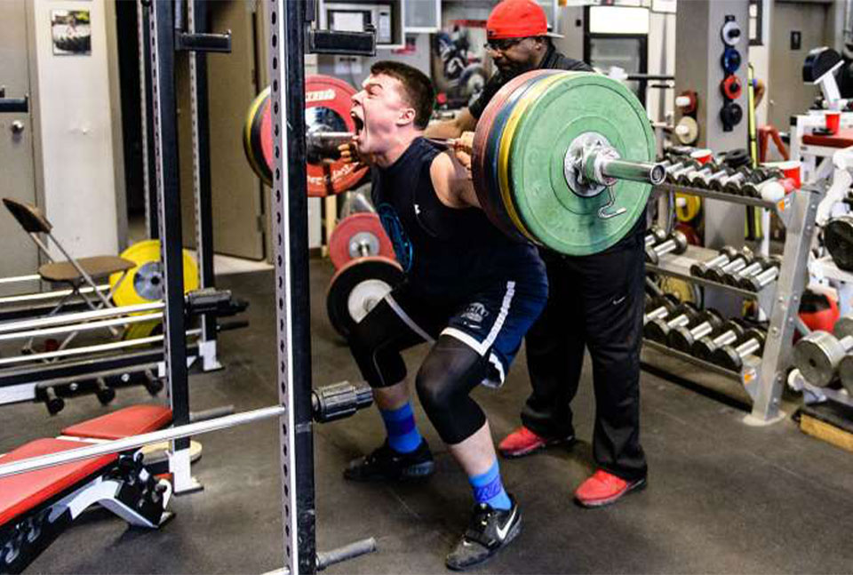 An athlete performing back squat with coach Clance Laylor spotting.