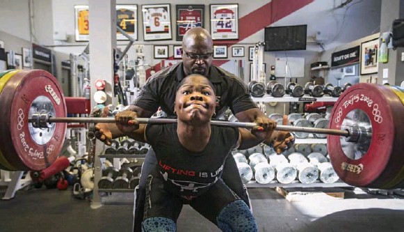 Maya Laylor performing back squat with her dad Clance Laylor spotting behind her.